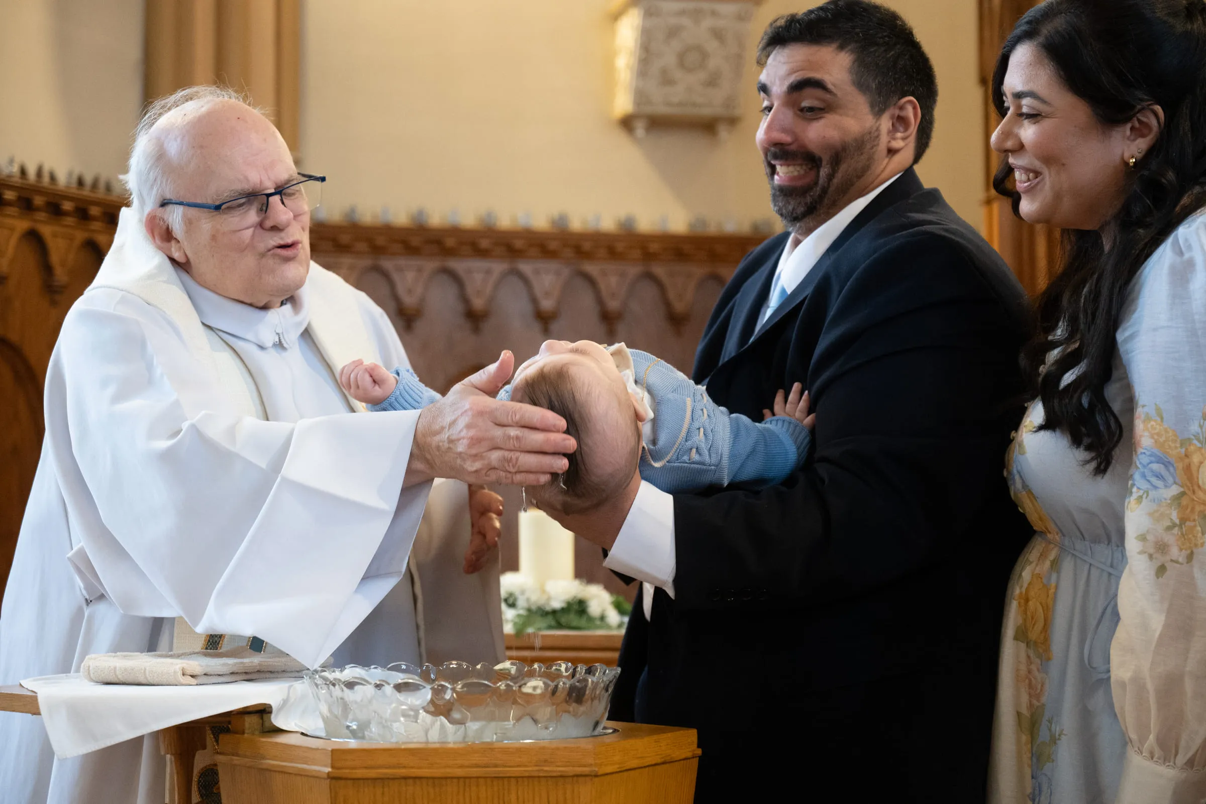 The priest pouring water over the baby’s head at the font as his parents look on