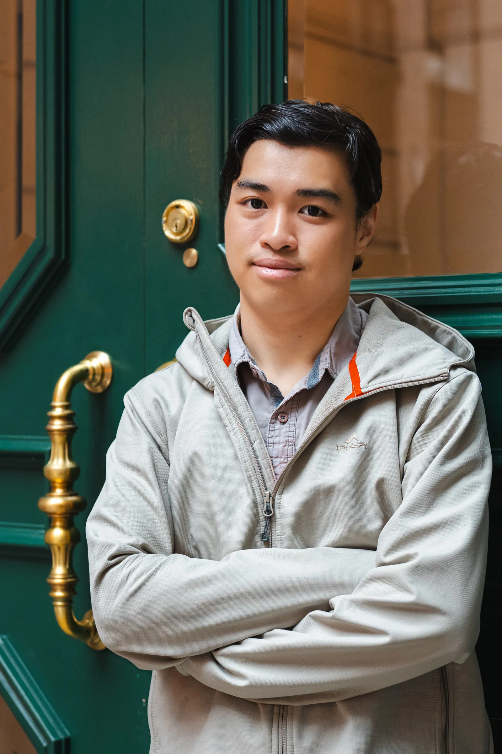 A young man standing with folded arms in front of a dark green door with a brass handle